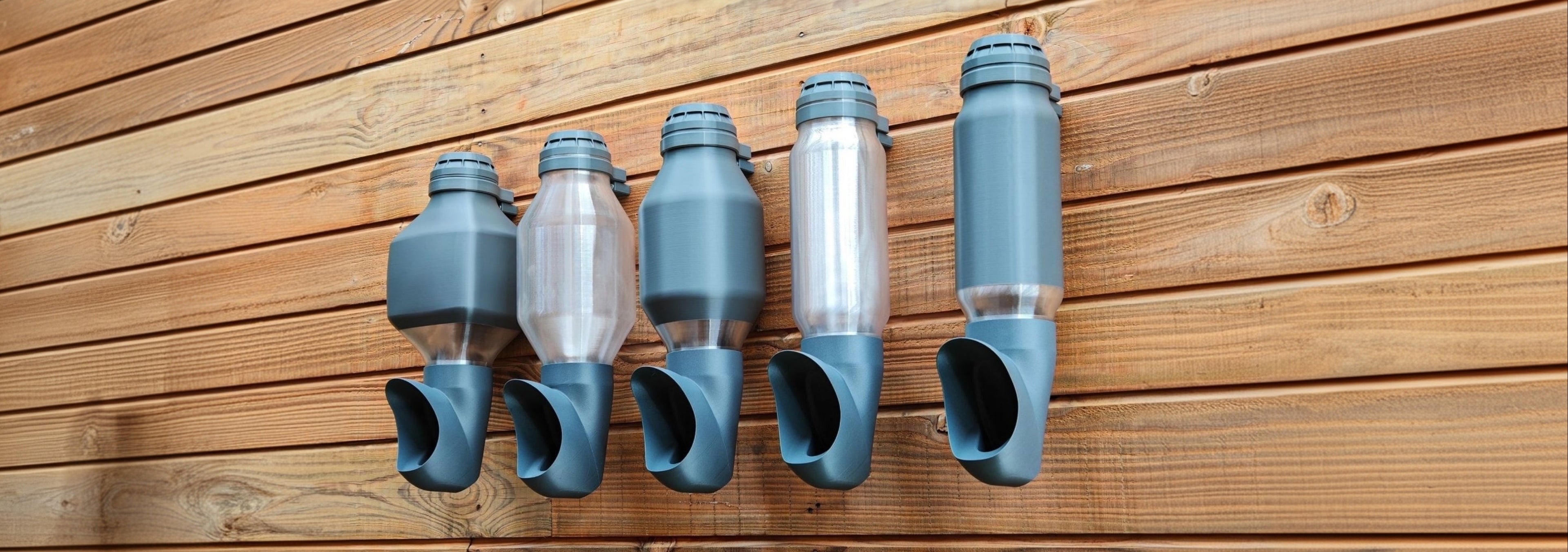 Photo de plusieurs mangeoires suspendues Cocobol pour poules, réservoirs transparents et opaques, fixées sur un mur en bois.