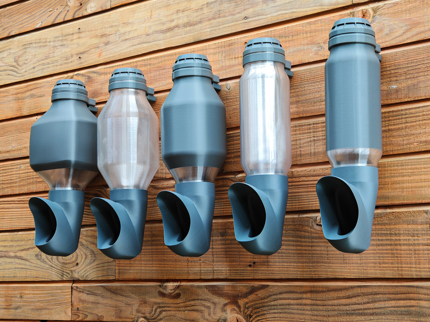 Photo de plusieurs mangeoires suspendues Cocobol pour poules, réservoirs transparents et opaques, fixées sur un mur en bois.