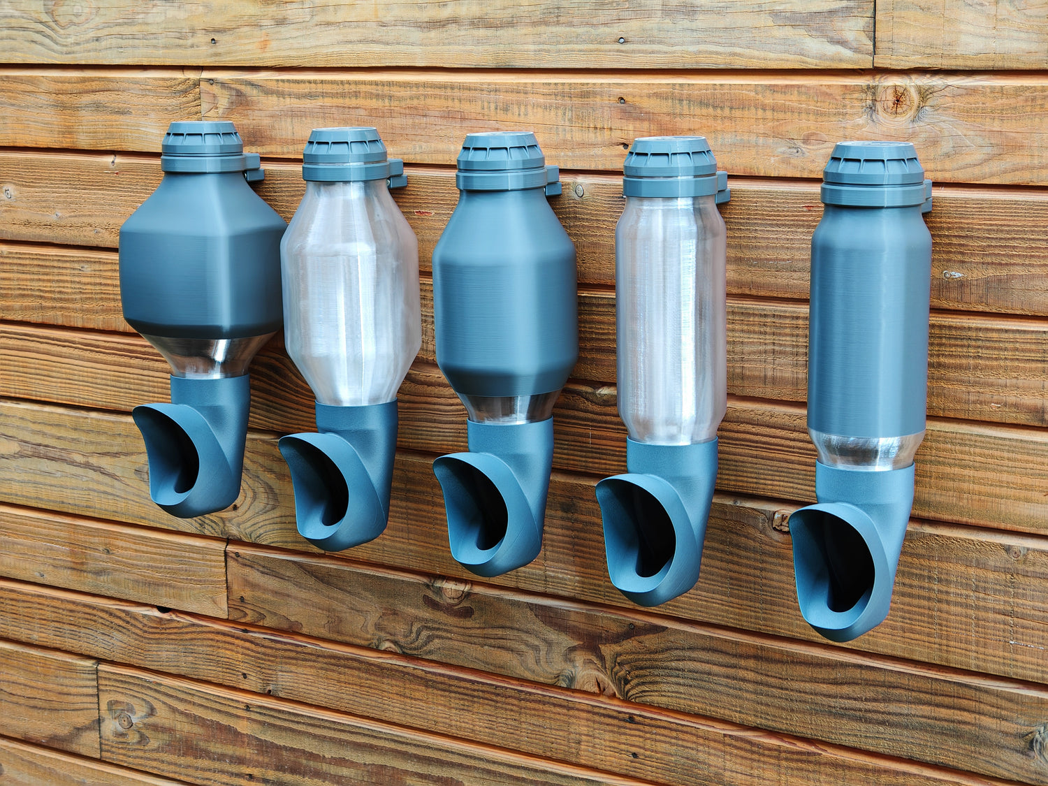 Photo de plusieurs mangeoires suspendues Cocobol pour poules, réservoirs transparents et opaques, fixées sur un mur en bois.